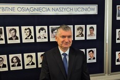 Dr hab. inż. Mariusz Korkosz, prof. uczelni na tle tablicy z fotografiami uczniów którzy byli laureatami Olimpiady Elektrycznej i Elektronicznej,